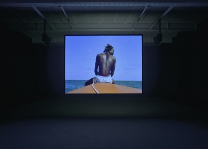 A dark room is illuminated by a projection of a man sitting on the stern of a boat.