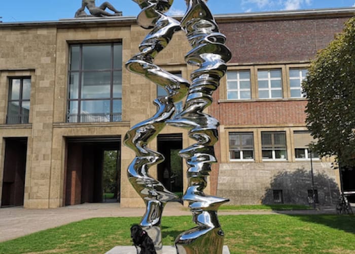 Sculpture in garden: two wavy silver pillars zig zag in front of a plain brown building.