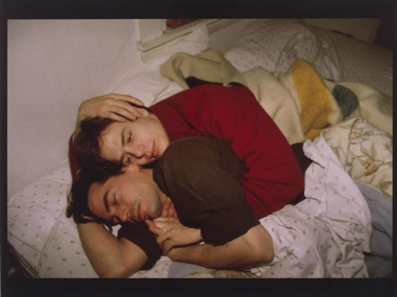 Image of two men wearing sweaters embracing in bed.