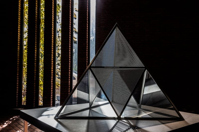 Installation view of a partially perforated, triangular metal structure in a darkened room with dramatic lighting.
