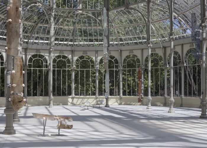 An outdoor, glass pavilion with archways features a vertical, tubular sculpture by Nairy Baghramian.