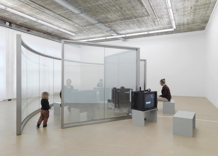 Multiple curved and straight glass panels are assembled next to TV's as multiple figures fill the bright space.