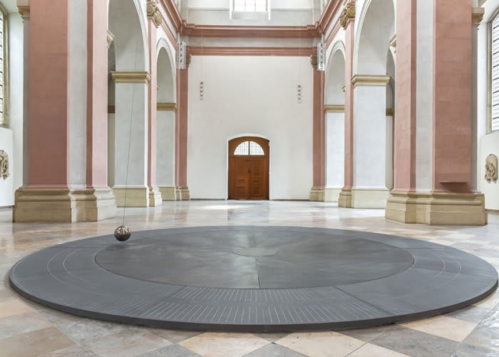 Image of a flat black disc on the ground of a large pink and white marble room with a metal ball attached to a string dangling above.