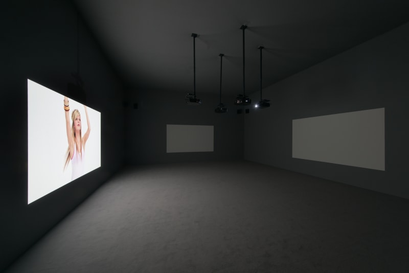 A dark gray room with 3 projection screens; on the left screen a girl dances dressed in white.