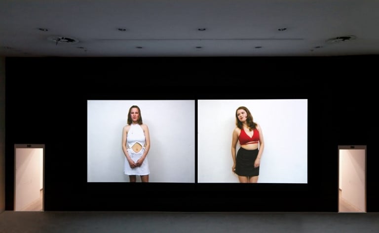 A dark room is illuminated by a video diptych of two young women speaking against a white wall.