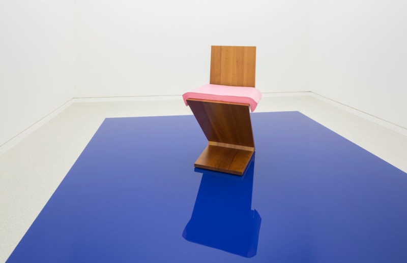 Geometric wooden sculpture with pink accent rests on reflective blue surface.