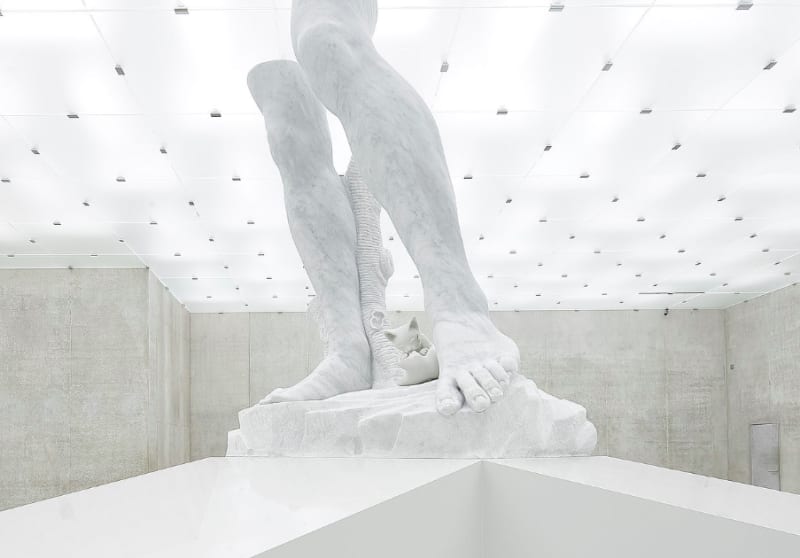 Detail image of a white marble sculpture of the legs of a man in a brightly illuminated white room.