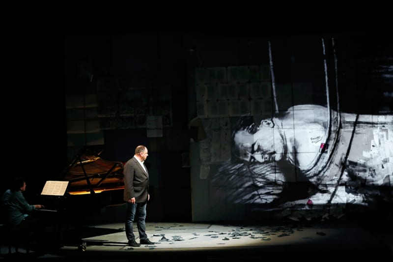 Person on stage under a spotlight looks to projection of larger-than-life black and white rendered person sleeping. To the left, a person plays the piano.