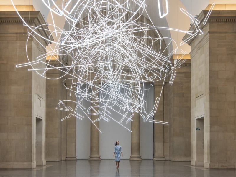 A large marble room features a hanging neon sculpture composed of long, thin and bending lines.