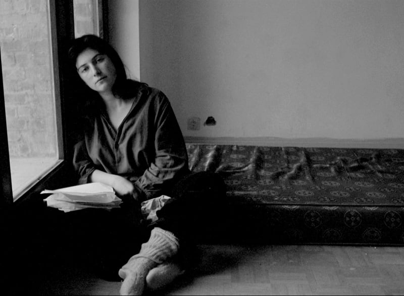 Black and white portrait of a person leaning against a window. They gaze into the camera. There is a mattress on the floor behind them.