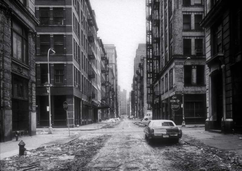 Black and white image of street with an old fashioned car parked on the curb.