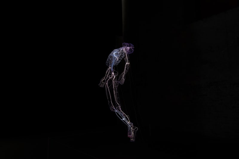 An image containing a skeletal human figure by Tavares Strachan, made of blue neon, purple neon, pyrex, transformers, and a MDF box.