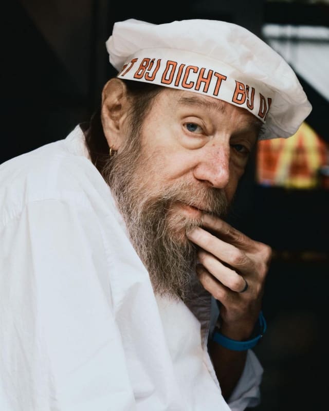 An image containing a portrait of Lawrence Weiner, wearing a hat with his art
