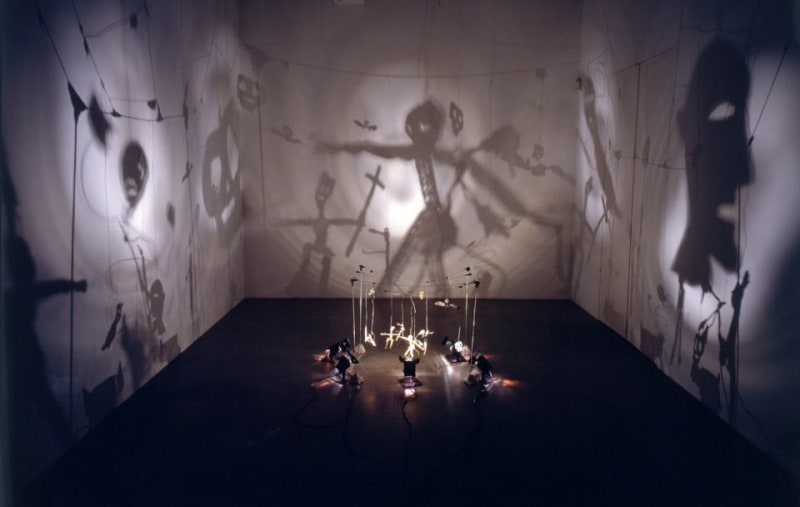Installation of illuminated suspended dolls in center of room that create shadow silhouettes on the walls.