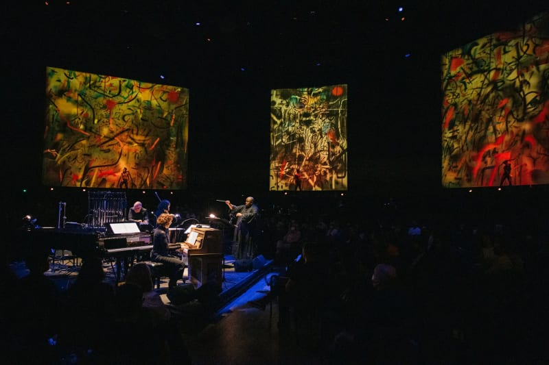 The composer Tyshawn Sorey, center, conducting his “Monochromatic Light (Afterlife)” at the Park Avenue Armory, where illuminated reproductions of paintings by Julie Mehretu surrounded the audience. Credit: Jeenah Moon for The New York Times
