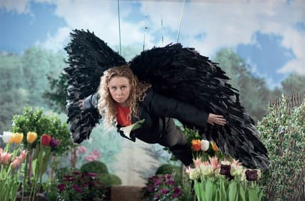 Person adorned with large black wings is suspended in front of a nature mural. Real flowers line the indoor floor.