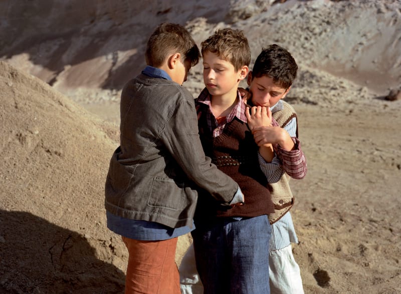 Three children huddle together with their eyes closed. Sand dunes surround them,