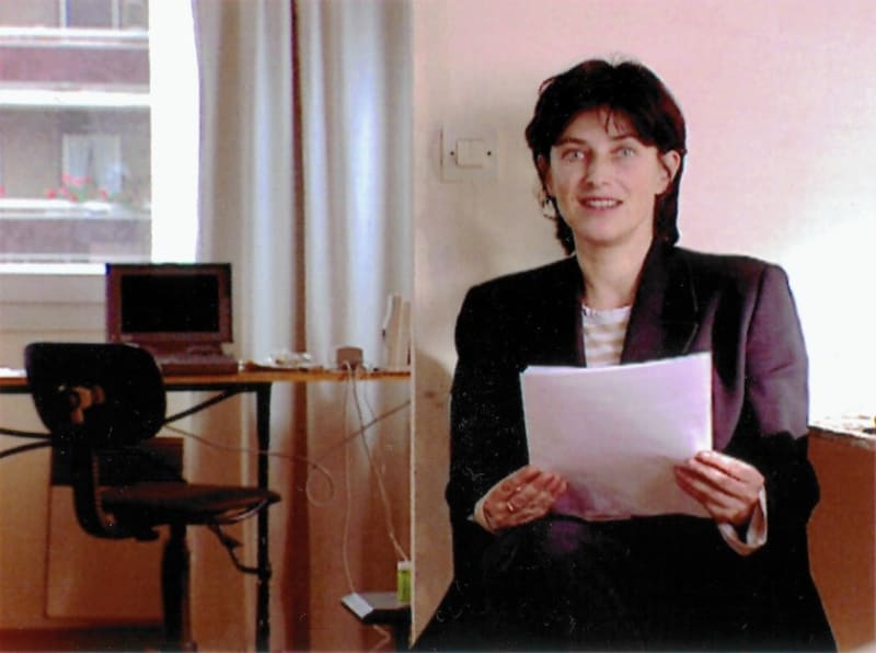Seated person holding paper looks into camera. To the left, a desktop computer.