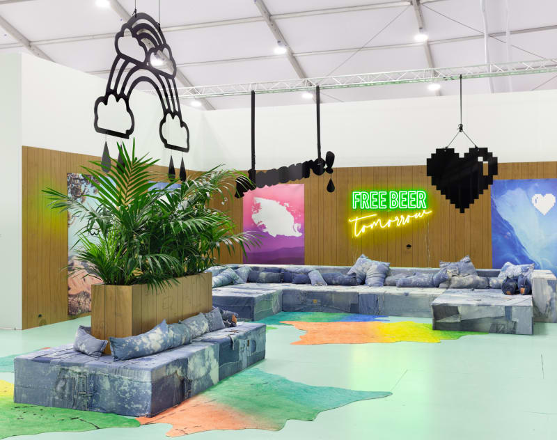 Wendy White. Free Beer Tomorrow, 2019. Hand- carved wood paneling, repurposed distressed denim, upholstery foam and wood custom furniture, digital prints on neoprene, hand painted faux calfskin rugs, paper cigarettes, custom floor paint, LED sign, live plants, Dimensions variable