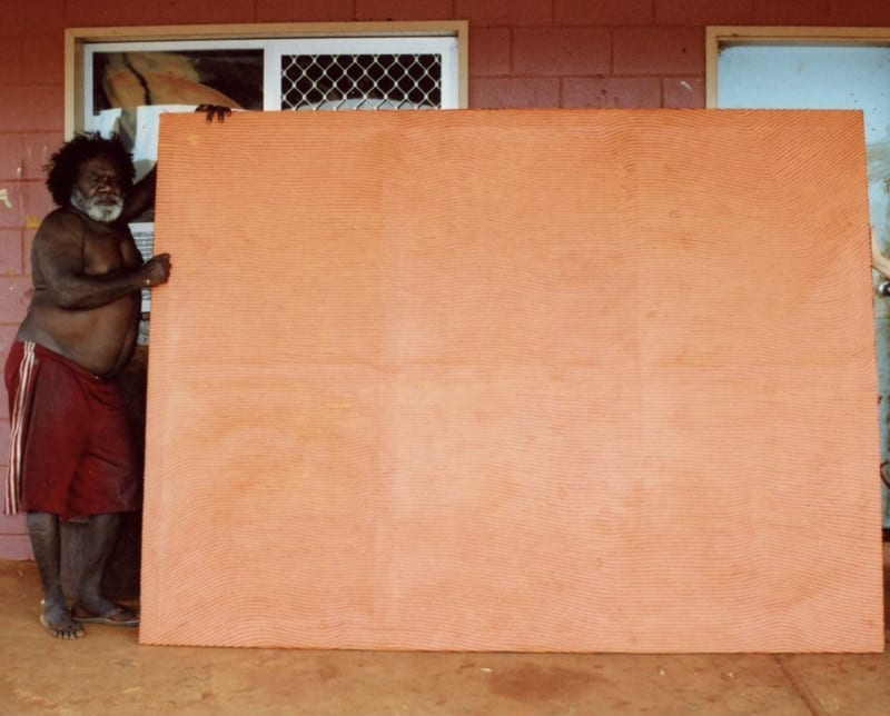 Photograph of George Tjungurrayi, seen holding one of his paintings distinguished by his signature abstract use of optical stripes