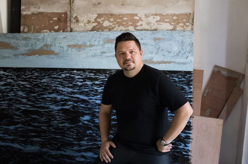 Portrait photograph of a man in his mid-thirties with cropped dark hair and a goatee. He wears a black T-shirt and stands in front of a large painting of a tranquil sea. 