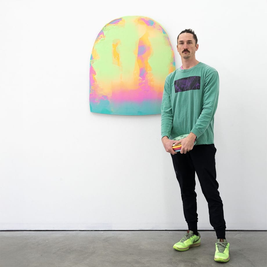 Artist Nick Schleicher stands next to an original abstract painting. The canvas is dome shaped and painted a wash of neon and electric greens yellows and blues