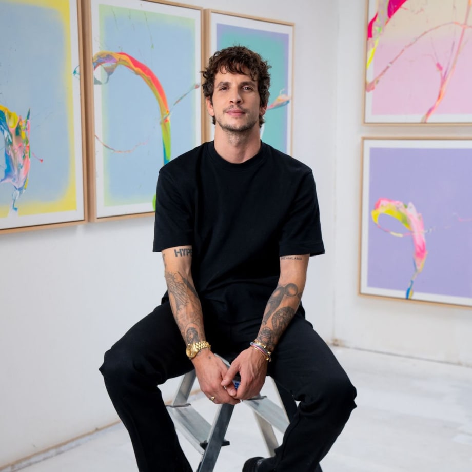 Photo of artist Cevin Parker seated in front of multiple colorful artworks with pastel and rainbow paint splatter hanging behind him