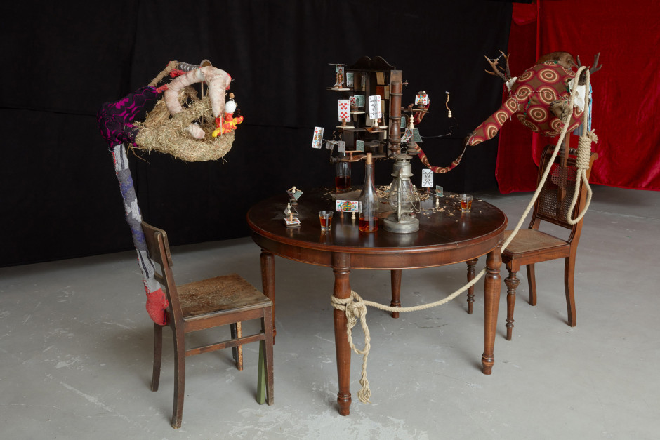 Skat Chess, 2017  wooden chairs, wooden table, wood, chess piece, whiskey, bottle, glasses, play dough, socks, deer antlers, stuffed muskrat, straw, egg shells, fabric, buttons  151 x 222 x 119 cm  59 ½ x 87 ⅜ x 46 ⅞ in.