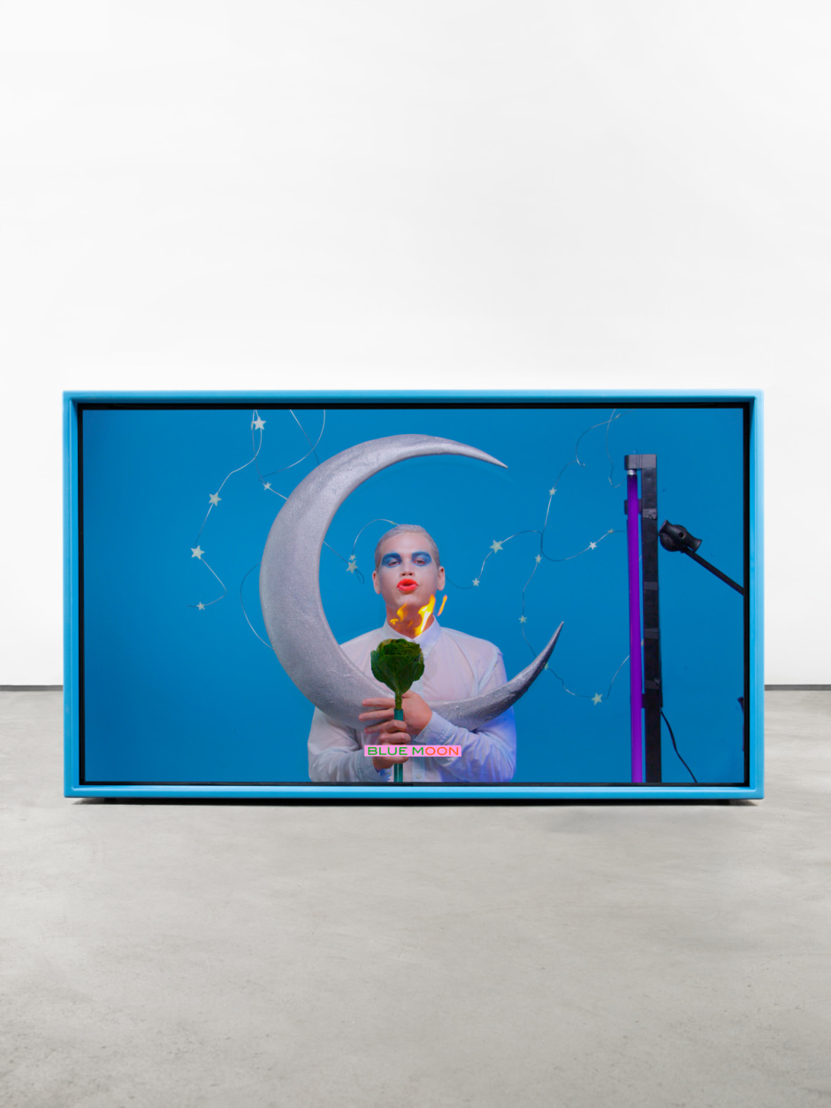 Alex Da Corte  Blue Moon, 2017  video, color/sound  duration: 2 min 45 sec  Blue Moon, 2017, is a theatrical video portrait by Alex Da Corte, originally commissioned for the digital art exhibition Midnight Moment presented by Times Square Arts in partnership with the Whitney Museum of American Art at Times Square, New York, 2017.  The short video features an actor dressed in exaggerated stage make up, slowly crooning along to the popular 1934 Richard Rodgers and Lorenz Hart ballad. The actor holds a silver crescent moon and is surrounded by a constellation of cut-out stars, conjuring a surreal, cosmic karaoke as the lyrics light up the bottom of the screen.  Celestial forms act as a recurring motif within the artist’s work – most recently in his Horoscope series and As Long as the Sun Lasts, the 2021 Met Roof Garden Commission – as symbol for romance, aspiration and melancholia. Here, their flattened forms recall commercial signage and theatrical stage settings, both seductive and surreal.  In Blue Moon Da Corte reflects upon the enduring cultural fascination with finding love, as depicted in popular and commercial entertainment, conjuring a simultaneously wry and romantic vision of this search. The presentation mirrors this exploration of the commercialised representation of human experience. The work is displayed as a video sculpture, encased within an electric blue steel structure designed by the artist; its abstracted modular form resonating both with the art historical legacy of American Minimalist installation and subtly redolent of box-like family TVs that emerged within the song’s era.  The sing-a-long presentation likewise encourages the audience to participate in a collective experience. Of which the artist has stated: ‘The karaoke stage is a perfect melting pot for the good and the bad singers of the world to be what they want to be or what they don’t want to be without recourse. It is a safe space.’